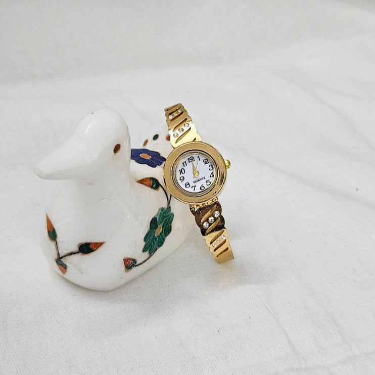 A gold-tone women's watch with a white dial and patterned brown strap, displayed on a white ceramic duck figurine against a white fabric background.